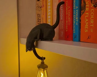 May include: A black cat figurine perched on the edge of a shelf, featuring a long, curved tail that holds a small, gold-colored lantern. The lantern emits a warm, glowing yellow light, illuminating the space below the shelf in a soft, ambient way.