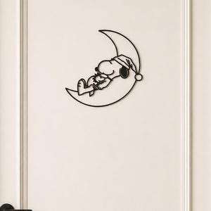 Könnte beinhalten: Schwarze Metall-Silhouette von Snoopy, der auf einem Halbmond schläft und eine Schlafhaube trägt. Das Kunstwerk ist an einer weißen Tür angebracht. Die Tür hat einen schwarzen Griff.