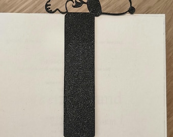 May include: A black metal bookmark featuring a Snoopy design, resting on the corner of a book. The bookmark has a rectangular body with a Snoopy silhouette at the top. The bookmark is made of a textured material.
