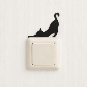 Op de afbeelding: Een zwart silhouet van een kat in een uitgerekte houding, geplaatst direct boven een vierkante, gebroken witte lichtschakelaar. De katfiguur is plat en effen zwart, waarbij het dier met de voorpoten naar voren gestrekt en de staart omhoog staat.