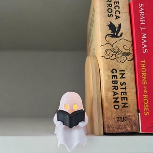 May include: A small, white, translucent ghost figurine holding a black book, positioned next to a wooden bookend and two books titled In Steen Gebrand by Rebecca Yarros and Thorns and Roses by Sarah J. Maas, with glowing yellow eyes.