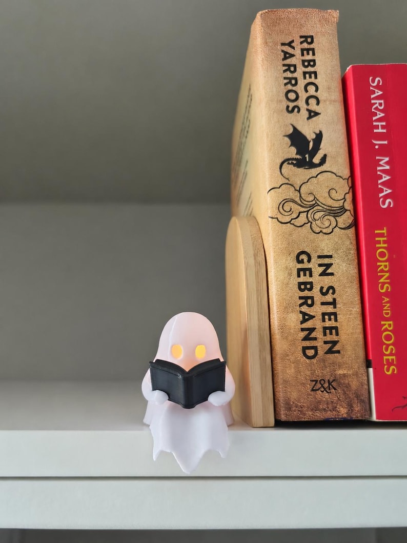 May include: A small, white, translucent ghost figurine holding a black book, positioned next to a wooden bookend and two books titled In Steen Gebrand by Rebecca Yarros and Thorns and Roses by Sarah J. Maas, with glowing yellow eyes.