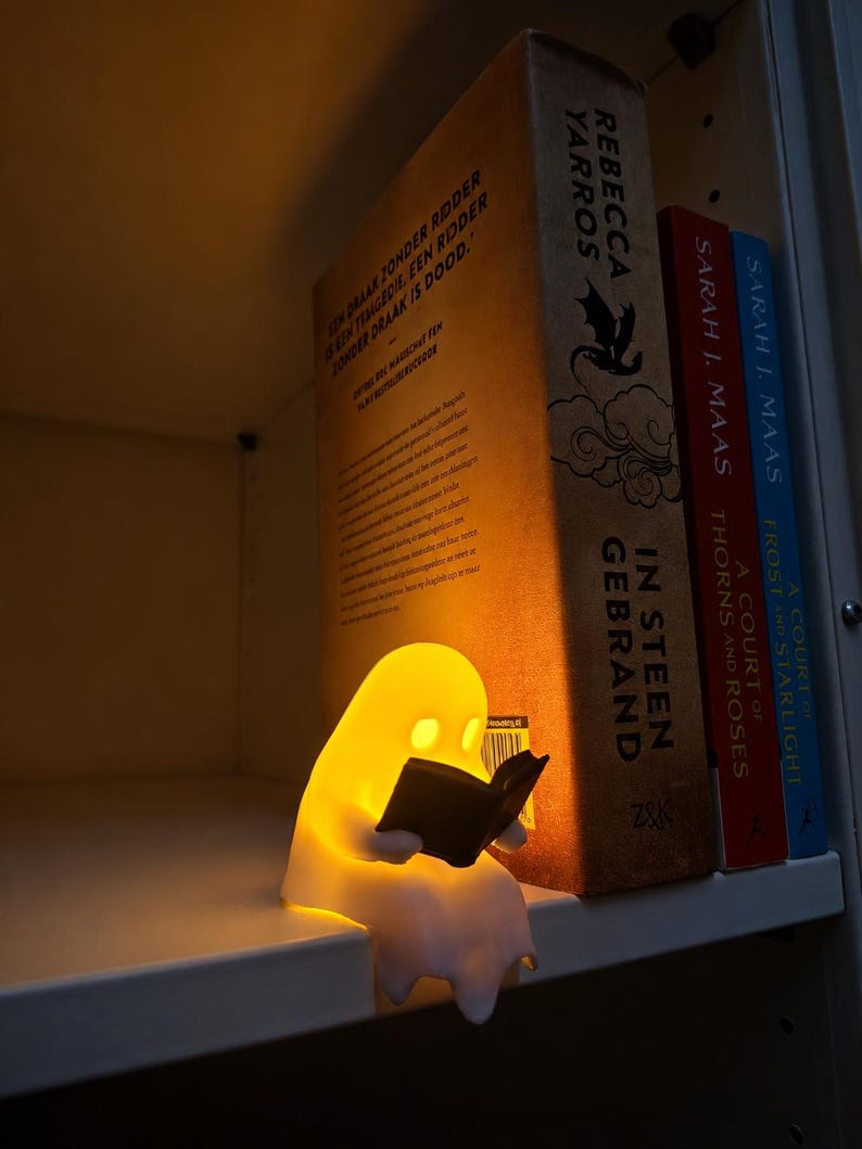 May include: A small, glowing, white ghost-shaped lamp sits on a shelf, holding a miniature black book. Behind the lamp are three books: "In Steen Gebrand" by Rebecca Yarros, "A Court of Thorns and Roses" by Sarah J. Maas, and "A Court of Frost and Starlight".