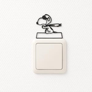 Może przedstawiać: Biały, kwadratowy włącznik światła z czarną sylwetką z drutu przedstawiającą postać z kreskówki Snoopy w okularach przeciwsłonecznych i szaliku, umieszczoną na płytce przełącznika jako dekoracyjny akcent ścienny.
