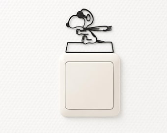 May include: A white square light switch cover featuring a black wire-frame silhouette of the cartoon character Snoopy wearing sunglasses and a scarf, positioned on top of the switch plate as a decorative wall accent for home interiors.