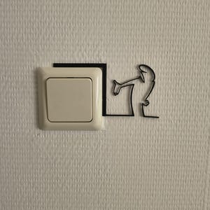 Op de afbeelding: Een vierkante, gebroken witte lichtschakelaar aan een muur, voorzien van een zwarte, dunne lijnsilhouetsticker van een figuur die over de bovenrand van de schakelaar leunt en met het mechanisme lijkt te interageren via een doorlopend lijnontwerp.