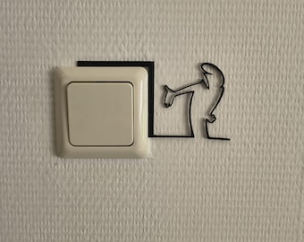 May include: A square, off-white light switch mounted on a wall, featuring a black, thin-lined silhouette decal of a figure leaning over the top edge of the switch, appearing to interact with the mechanism through a minimalist, continuous line design.