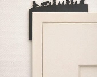 May include: A black metal door topper featuring a silhouette of a procession of figures. The topper is mounted on the corner of a white door. The figures appear to be walking across a hill.