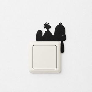 Könnte beinhalten: Ein mattschwarzer dekorativer Wandaufkleber mit der Silhouette eines Hundes und eines kleinen Vogels, der auf seinem Kopf sitzt, direkt über einem quadratischen weißen Lichtschalter angebracht. Das Design ist flach und minimalistisch.