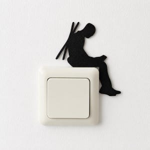 Può includere: Un adesivo a silhouette nera raffigurante una persona seduta che legge un libro, posizionato sull'angolo superiore destro di un interruttore della luce quadrato color bianco sporco. La figura ha due sottili linee verticali sulla schiena.
