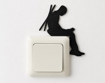 May include: A black silhouette sticker of a person sitting and reading a book, positioned on the top right corner of a square, off-white light switch. The figure has two thin vertical lines extending from the back, resembling skis or poles.