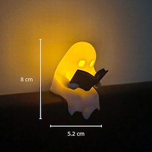 May include: A white ghost-shaped night light emitting a warm yellow glow, holding a small black book. The figure measures 8 cm in height and 5.2 cm in width, as indicated by the white measurement lines and text labels visible in the image.