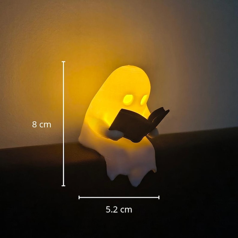 May include: A white ghost-shaped night light emitting a warm yellow glow, holding a small black book. The figure measures 8 cm in height and 5.2 cm in width, as indicated by the white measurement lines and text labels visible in the image.