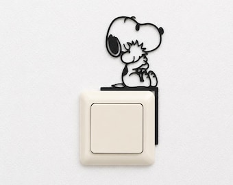 May include: A black metal wall decal featuring the cartoon character Snoopy hugging Woodstock, positioned directly above a square, off-white light switch. The cutout design is thin and outlines the two characters in a simple, flat silhouette.