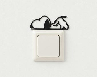 May include: A black silhouette decal of a cartoon dog lying down, positioned directly above a square white light switch. The decal features the distinct outline of the character Snoopy resting with ears and paws visible in a flat, matte finish.