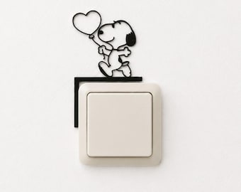May include: A black metal wall decal featuring a cartoon dog character holding a heart-shaped balloon, positioned above a square, off-white light switch. The thin, wire-like silhouette of the character is mounted directly onto the wall surface.