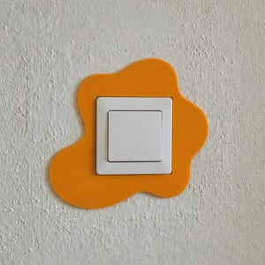 Può includere: Un interruttore della luce quadrato bianco montato su una parete, incorniciato da una piastra arancione di forma organica irregolare. La cornice arancione presenta bordi lisci e arrotondati e una superficie piatta e uniforme.
