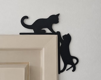 May include: A black metal wall decoration featuring two cat silhouettes positioned at the corner of a door frame. One cat is depicted walking horizontally along the top edge, while the second cat is shown climbing vertically down the side frame.