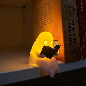 Peut inclure: Une petite lampe blanche en forme de fantôme posée sur une étagère. La figurine est faite d'un matériau translucide qui émet une lumière jaune chaude et tient un petit livre noir ouvert dans ses mains, semblant le lire avec attention.