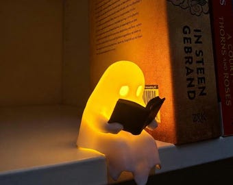 May include: A small, glowing, white ghost-shaped lamp sitting on a shelf. The ghost figure is made of a translucent material that emits a warm yellow light and is holding a small, black, open book in its hands, appearing to read it intently.