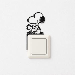Pode incluir: Um adesivo de parede de metal preto com os personagens de desenho animado Snoopy e Woodstock sentados em cima de um interruptor de luz quadrado branco. O design é uma silhueta fina e plana que se fixa no canto da placa do interruptor.