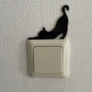 Op de afbeelding: Een zwart silhouet van een kat in een uitrekkende houding, direct boven een vierkante, gebroken witte lichtschakelaar geplaatst. De platte kattenfiguur heeft een gebogen staart en uitgestrekte voorpoten als decoratief accent voor de schakelaar.