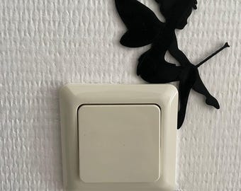 May include: A black silhouette of a fairy with wings and a wand positioned next to a square, off-white plastic light switch. The fairy figure is flat and appears to be attached to the wall surface adjacent to the light switch cover plate.
