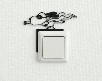 May include: A black metal wall decal featuring a cartoon dog wearing goggles and a flowing cape, positioned directly above a square white light switch. The silhouette design depicts the character in a flying pose, mounted on the wall surface.