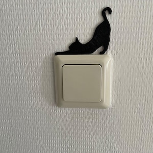 Könnte beinhalten: Eine schwarze, katzenförmige Lichtschalterabdeckung erstreckt sich über einen cremefarbenen Lichtschalter. Die Katze befindet sich in einer spielerischen Pose, mit nach oben gebogener Schwanzspitze. Der Hintergrund ist eine strukturierte weiße Wand.