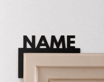 May include: A black metal door corner sign featuring the word NAME in bold, uppercase, sans-serif capital letters. The L-shaped design is mounted on the top corner of a light beige door frame, creating a clean and modern visual accent.