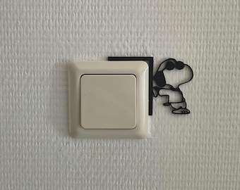 May include: A black, thin, wire-like decorative wall decal shaped like a cartoon dog wearing sunglasses, positioned to the right of a square, off-white plastic light switch. The decal features a corner bracket design that frames the light switch.