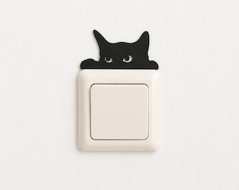 May include: A matte black cat silhouette decal positioned above a square, off-white light switch. The cat design features two pointed ears and two eyes peering over the top edge of the switch plate, creating a decorative accent for the wall.