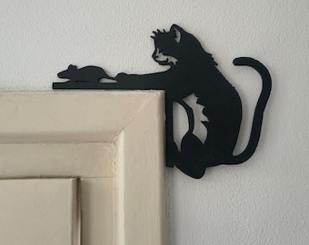 May include: A black silhouette wall decoration designed to fit on the corner of a door frame. The piece features a cat reaching out with one paw toward a small mouse positioned on the horizontal edge of the frame, creating a playful interaction.