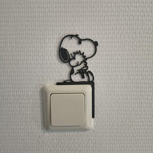 Könnte beinhalten: Eine schwarze Snoopy-förmige Lichtschalterabdeckung ist über einem weißen Lichtschalter angebracht. Die Abdeckung ist eine Silhouette von Snoopy, der sitzt und nach oben schaut. Der Lichtschalter ist quadratisch mit einem rechteckigen Knopf.