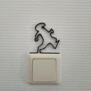 Könnte beinhalten: Schwarze Metall-Silhouette einer rennenden Person, montiert über einem Lichtschalter. Der Lichtschalter ist cremefarben und rechteckig, vor einer strukturierten weißen Wand. Das Kunstwerk verleiht dem Raum eine spielerische Note.