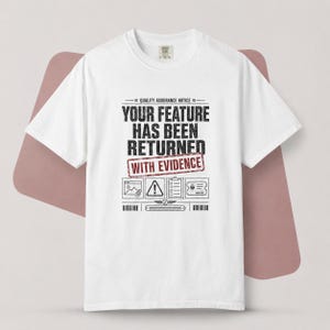 Op de afbeelding: Een wit t-shirt met korte mouwen en zwarte tekst die QUALITY ASSURANCE NOTICE YOUR FEATURE HAS BEEN RETURNED WITH EVIDENCE in een rood kader luidt, samen met iconen van een document, waarschuwingsbord, klembord en een ticket met een kever.