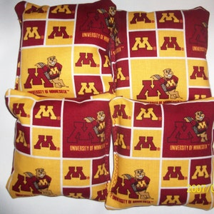 May include: Set of six bean bags featuring the University of Minnesota Golden Gophers logo. The bean bags are made of a red and gold fabric with a white background. The logo features a gopher holding a football.