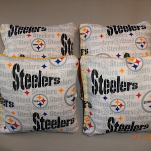 May include: Four white bean bags with a black and yellow Steelers logo and the word "Steelers" repeated on the fabric. The bean bags have a yellow trim.