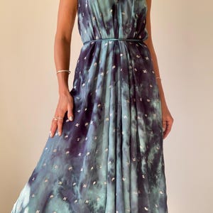 May include: A flowing, tie-dye maxi dress in shades of teal and navy blue, adorned with silver metallic accents. The dress features a halter neckline and a cinched waist, creating an elegant silhouette. The fabric drapes gracefully, suggesting a lightweight material.