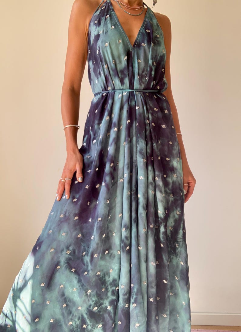 May include: A flowing, tie-dye maxi dress in shades of teal and navy blue, adorned with silver metallic accents. The dress features a halter neckline and a cinched waist, creating an elegant silhouette. The fabric drapes gracefully, suggesting a lightweight material.