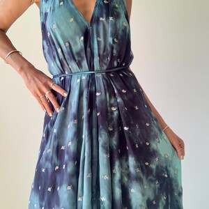 May include: A halter-neck dress in shades of teal and navy blue, featuring a tie-dye pattern with silver accents. The dress has a V-neckline and a flowing, pleated skirt. A thin belt cinches the waist. The dress is made of a lightweight fabric.