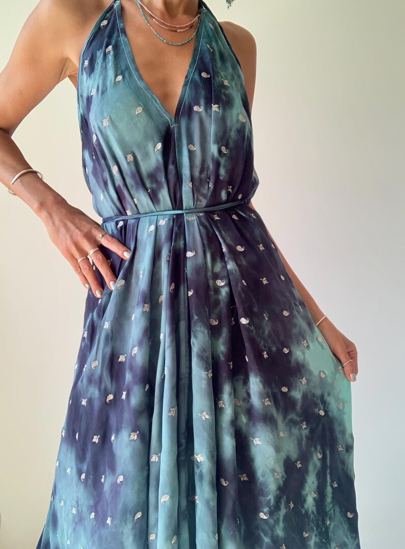 May include: A halter-neck dress in shades of teal and navy blue, featuring a tie-dye pattern with silver accents. The dress has a V-neckline and a flowing, pleated skirt. A thin belt cinches the waist. The dress is made of a lightweight fabric.