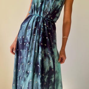 May include: A floor-length halter dress featuring a deep teal and navy blue tie-dye pattern with small silver metallic leaf-shaped accents. The garment has a plunging V-neckline, a gathered waist with a thin matching tie, and a flowing skirt.