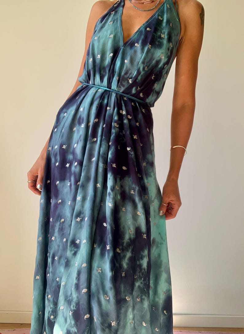 May include: A floor-length halter dress featuring a deep teal and navy blue tie-dye pattern with small silver metallic leaf-shaped accents. The garment has a plunging V-neckline, a gathered waist with a thin matching tie, and a flowing skirt.