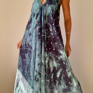 May include: A flowing, halter-neck maxi dress in a tie-dye pattern of teal and deep purple. The dress features a V-neckline and is embellished with small, silver-colored accents. The fabric drapes loosely, creating a relaxed silhouette.