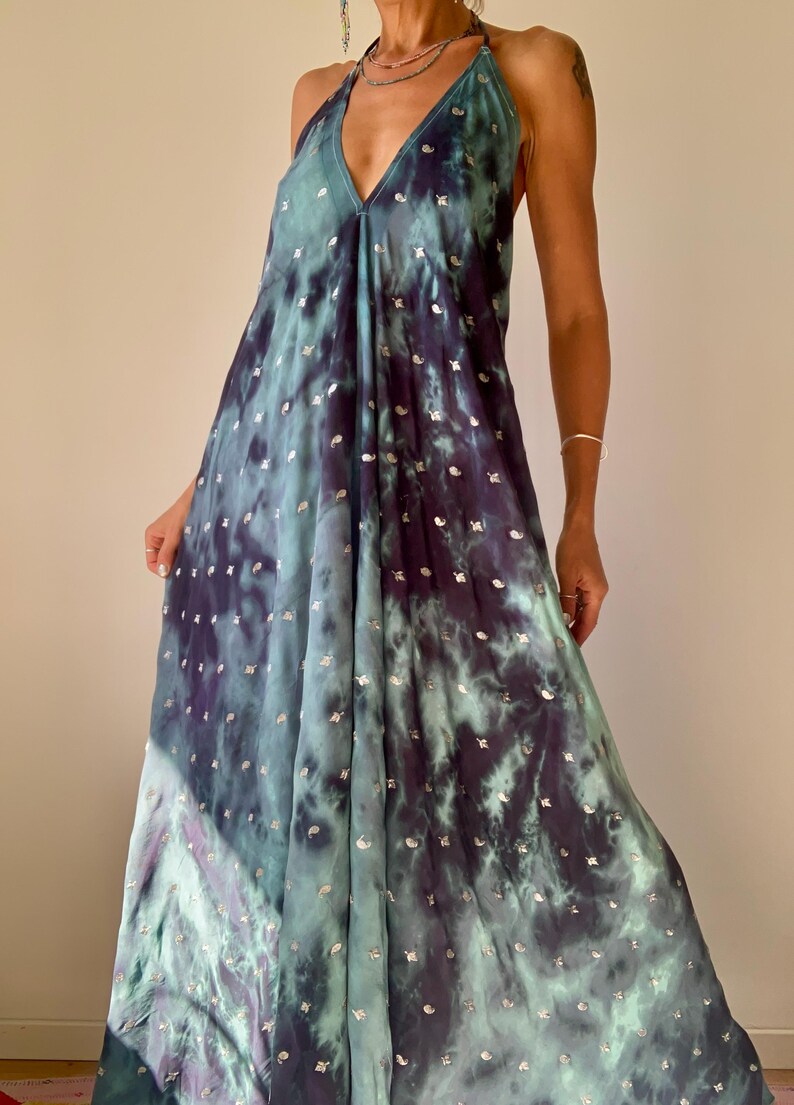 May include: A flowing, halter-neck maxi dress in a tie-dye pattern of teal and deep purple. The dress features a V-neckline and is embellished with small, silver-colored accents. The fabric drapes loosely, creating a relaxed silhouette.