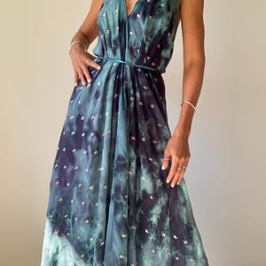 May include: A floor-length halter dress featuring a deep teal and navy blue tie-dye pattern with small, scattered metallic silver accents. The garment has a plunging V-neckline, a thin matching tie at the waist, and a flowing, loose silhouette.