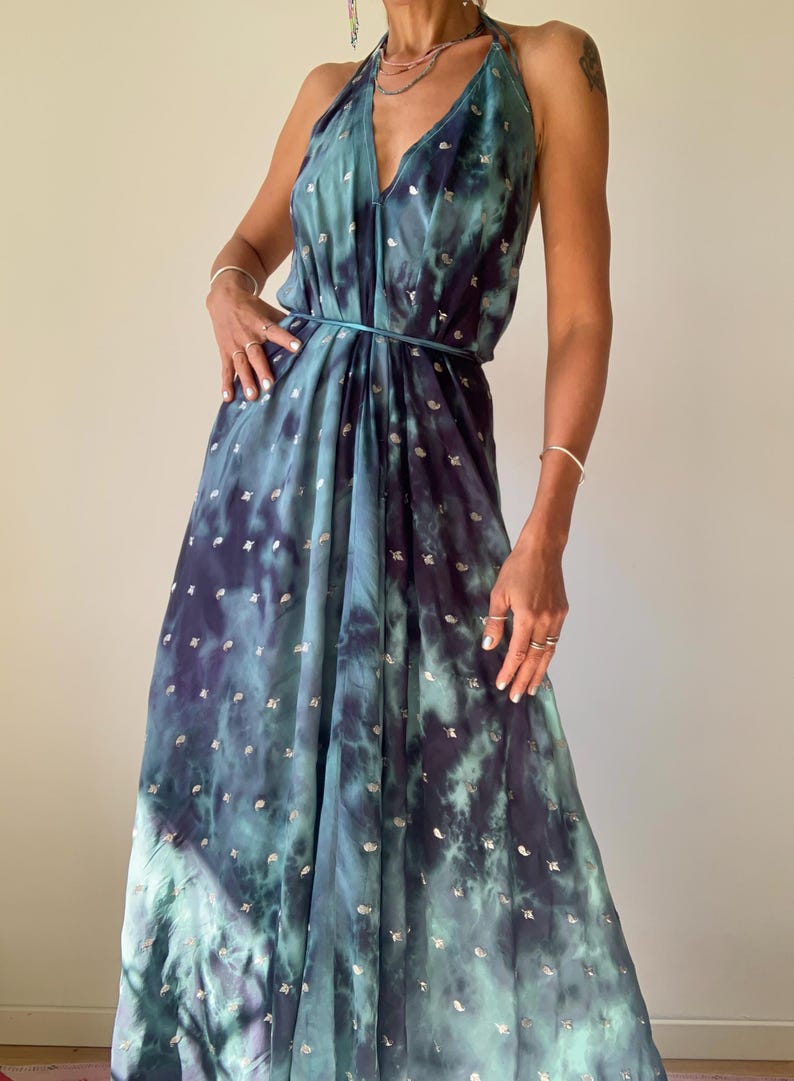 May include: A floor-length halter dress featuring a deep teal and navy blue tie-dye pattern with small, scattered metallic silver accents. The garment has a plunging V-neckline, a thin matching tie at the waist, and a flowing, loose silhouette.