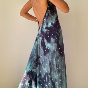 May include: A flowing, backless dress in shades of teal, purple, and navy, with a tie-dye effect. The fabric is adorned with small, silver-colored embellishments. The dress has a halter-style neckline and a long, elegant skirt.