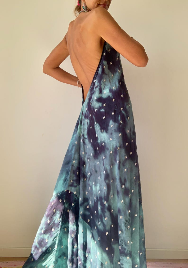 May include: A flowing, backless dress in shades of teal, purple, and navy, with a tie-dye effect. The fabric is adorned with small, silver-colored embellishments. The dress has a halter-style neckline and a long, elegant skirt.